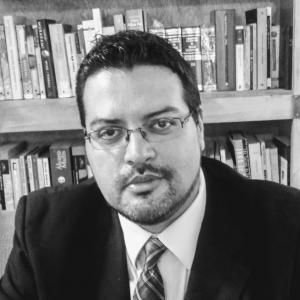  Aharon Aravizu Ramírez in glasses and a suit sits in front of a bookshelf filled with assorted books. His expression is serious, conveying a professional tone.