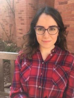 Ana Marqués García with long brown hair and glasses sits on a bench in front of a brick wall, wearing a red plaid shirt. She has a calm, pleasant expression.