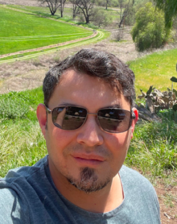 Ernesto Amaya in sunglasses takes a selfie outdoors, with a backdrop of lush green fields and trees under a clear blue sky, conveying a relaxed, sunny vibe.