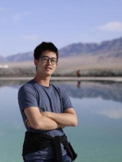 Rui Chu by a calm lake with mountains in the background under a clear blue sky. They appear relaxed and content, with arms crossed.
