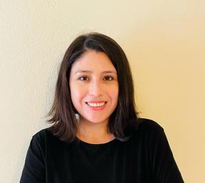 Bernadette Ramírez  with shoulder-length dark hair smiles warmly at the camera against a plain, light-colored background. They are wearing a black top.