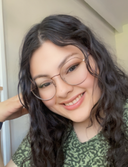 Carina Paredes Rivera smiling with long, wavy hair and glasses, wearing a green patterned top. She is indoors with soft lighting, creating a warm and cheerful atmosphere.