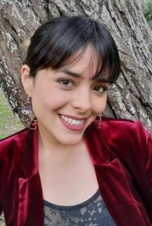 Carmen Araujo with short black hair, wearing a red velvet jacket, poses in front of a large tree trunk, conveying a warm, cheerful mood.