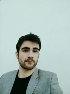 Salvador Del Ramo Zornoza with short dark hair and a beard wears a gray blazer over a black shirt. He stands against a plain off-white background, conveying a calm and composed expression.