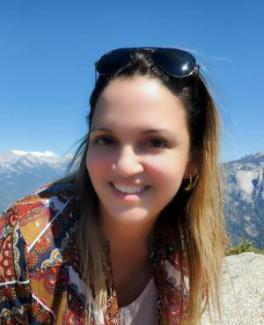 Carol Morejón smiling with sunglasses on head, wearing a patterned jacket, against a backdrop of mountains and clear blue sky, conveying a joyful and serene mood.